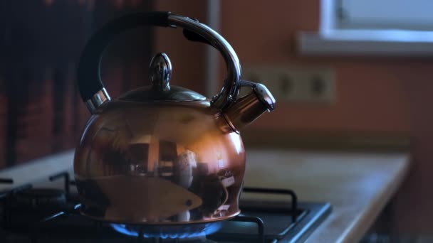 Gros plan d'une bouilloire à thé en acier inoxydable sur une cuisinière à gaz enflammée. Concept. Bouilloire chauffante avec le reflet de la cuisine sur sa surface métallique, préparation de boissons chaudes. — Video