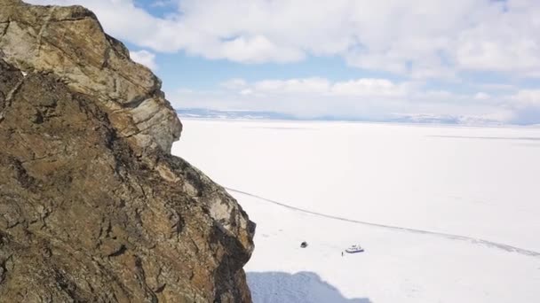 Vista aérea de um penhasco gigante localizado contra as montanhas no horizonte do lago congelado. Clipe. Vista do drone da rocha, água nevada do inverno do lago ou lagoa, e um grupo de pessoas que caminham perto do — Vídeo de Stock