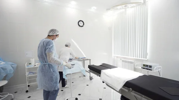 Médicos hombres y mujeres en la sala de procedimientos. Acción. Enfermera y cirujano preparando instrumentos y herramientas quirúrgicas, incluyendo bisturíes, pinzas y pinzas dispuestas sobre una mesa . —  Fotos de Stock