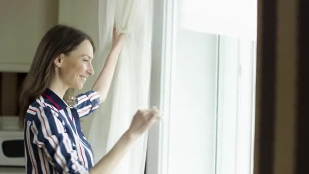 Tmavovlasá usměvavá žena otevře dveře balkónu a podívá se ven. Video. Boční pohled na šťastnou dámu doma dívající se ven oknem obývacího pokoje na slunný den. — Stock video