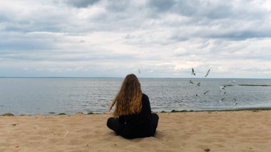 Finlandiya Körfezi, Saint Petersburg, Rusya 'daki bir kadının arka görüntüsü. Kavram. Siyah elbiseli, uzun saçlı, kumlu bir sahilde oturan ve uzaklara bakan genç bir kızın arka görüntüsü..