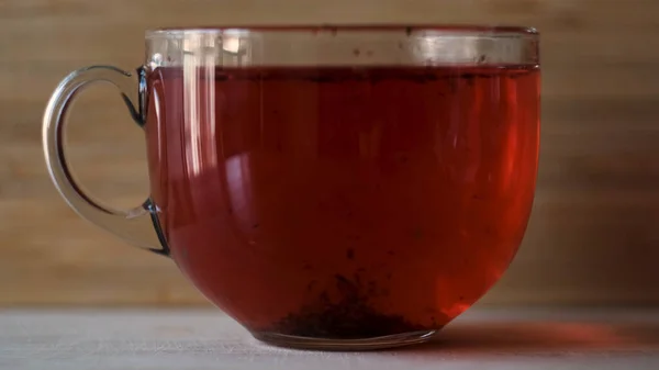 Gyümölcsvörös tea közeli felvétele egy átlátszó üvegbögrében, amely egy lapos asztal felületén áll. Fogantatás. Piros hibiszkusz tea fából készült falra háttér, koncepció egy alternatív módja annak, hogy kezelni és megelőzni influenza és — Stock Fotó