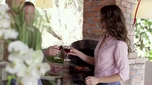 Hombre y mujer beben vino, hablando y sonriendo dentro de la cabaña. Vídeo. Vista lateral de un par de hombres y mujeres chating y flurting mientras que el hombre asando carne en la chimenea durante la fiesta de barbacoa. — Vídeos de Stock