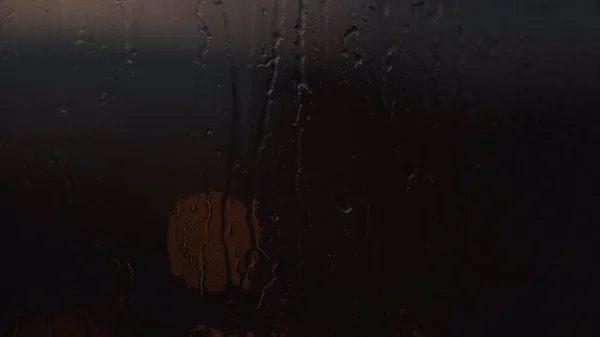 Drops of rain on window with one round light in the dark. Concept. Close up of glass wet surface with water droplets trickling down and the dim light of street lamp behind the window.