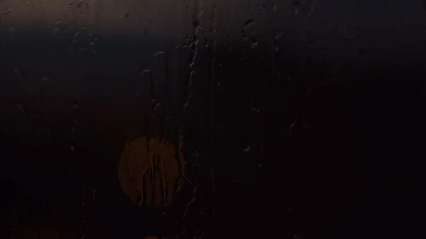 Esőcseppek az ablakban, egy kerek fénnyel a sötétben. Fogantatás. Az üveg nedves felületének közelsége vízcseppekkel és az ablak mögötti utcai lámpa halvány fényével. — Stock Fotó