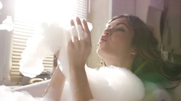 Jovem mulher atraente soprando espuma e brincando com ele. Vídeo. Sol brilhando através da janela no banheiro onde morena tomando banho de espuma e relaxante . — Fotografia de Stock