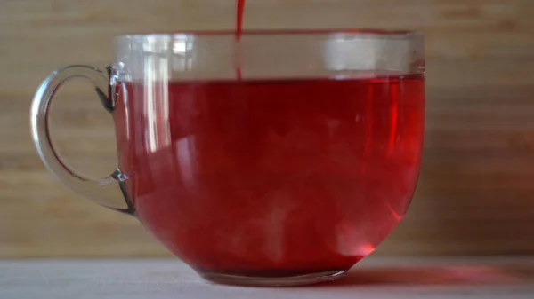 Doppa röd tepåse i en transparent mugg med varmt vatten på trävägg bakgrund. Begreppet. Närbild av bryggning av rött karkade te i en stor kopp med vatten. — Stockfoto