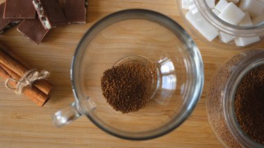 Bir kadının el yapımı granülatif kahve, şeker ve sıcak suyu bardağa koyarkenki görüntüsünü kapat. Kavram. Kahverengi ahşap yüzeyine çikolatalı şeffaf kahve yapmak ve...