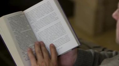 Evde kitap okuyan bir adamın omuzlarının üstünden. Video kaydı. Orta yaşlı erkek oturuyor ve kitap okuyor, sayfaları çeviriyor, evde dinleniyor..