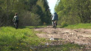 Köydeki bir köy yolunda bisiklet süren bir baba ve oğlunun arka görüntüsü. Hareket. Bir adam ve bir çocuk eğleniyor, yeşil ormanın yakınlarında spor yapıyorlar..