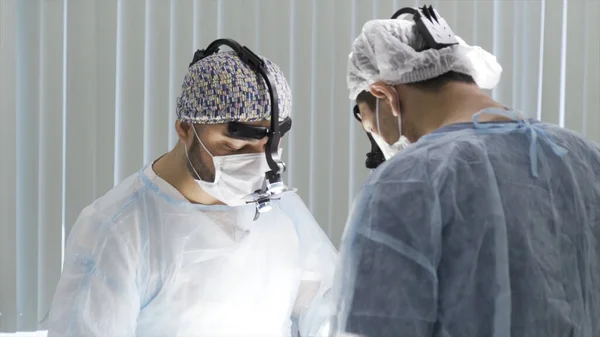 Two surgeons working at the operating clean room. Action. Two male doctors wearing sterile uniform and binocular loupe while performing surgery, concept of health care and medicine. — Stock Photo, Image