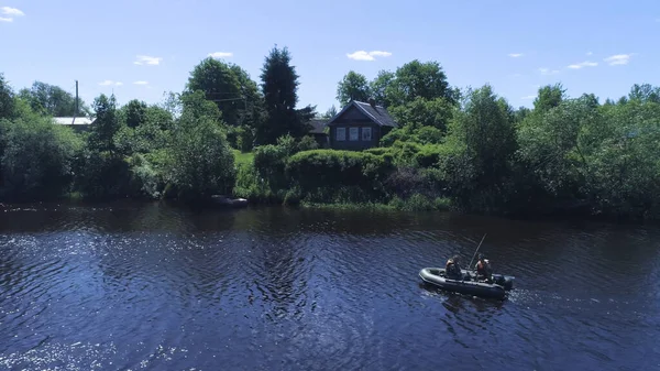 Рыбаки на надувной моторной лодке плавают по реке. Выстрел. Вид пожарных в лодке, плывущих по реке рядом с зеленым берегом с деревенскими домами. Летняя рыбалка на реке — стоковое фото