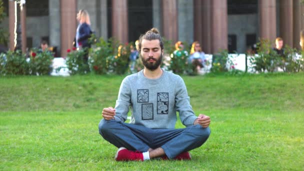 El hombre barbudo medita sobre hierba verde en el parque de la ciudad. Concepto. Elegante joven medita en la hierba verde en la bulliciosa ciudad. Hermoso hombre con estilo está interesado en la meditación — Vídeo de stock