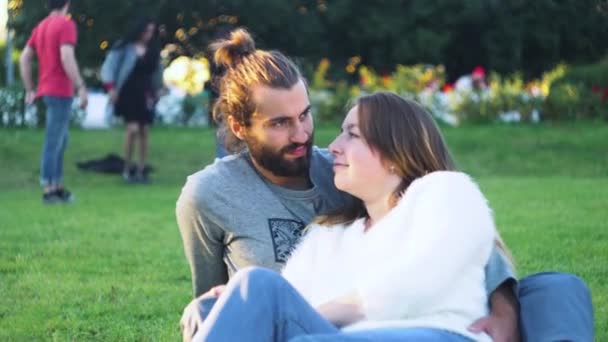 Красива пара пестить, сидячи на зеленій траві в парку. Поняття. Щаслива молода пара обіймається зеленою травою на сонці. Чоловік і жінка ніжно обіймаються і посміхаються сидячи на траві в парку — стокове відео