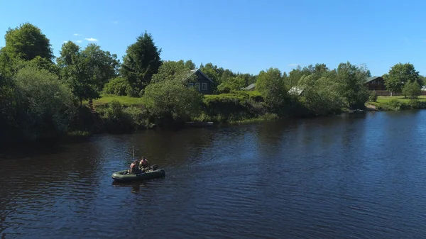 Ein Mann in einem Schlauchboot, das am Ufer des Flusses im Dorf rudert. Schuss. Das Konzept des Tourismus, Urlaub, Hobbys und Outdoor-Aktivitäten, zwei Männer in einem Boot in der Nähe von Holzhaus und grünen Bäumen segeln — Stockfoto
