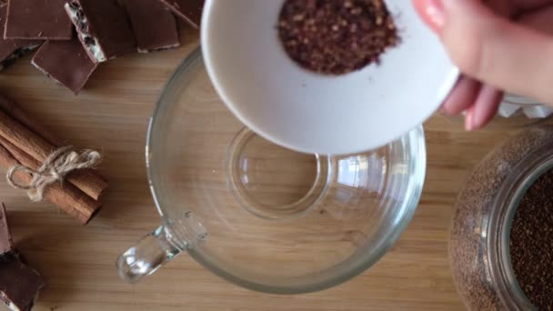Blisko góry widok wylewania suchego czerwonego hibiskusa liści herbaty w przezroczystym kubku. Koncepcja. Proces wytwarzania filiżanki czerwonej herbaty owocowej na drewnianym tle stołu z czekoladą i cynamonem. — Wideo stockowe