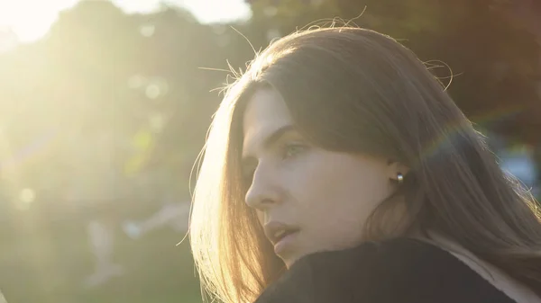 Perfil da jovem mulher bonita no fundo da luz solar. Mídia. Os raios de sol quentes caem no cabelo da bela mulher no parque. Mulher romântica endireita o cabelo no fundo do sol — Fotografia de Stock