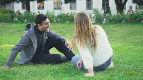 젊은 남녀는 공원의 풀 위에 앉아 웃는다. 미디어. 아름다운 부부는 푸른 풀 위에 앉아 긴장을 풀고 웃음을 터뜨린다. 잔디 위에서 웃는 학생 부부 — 스톡 사진