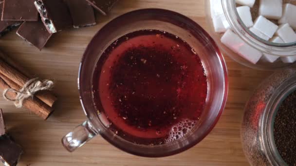 Process of brewing red karkade tea, close up top view. Concept. Cup of freshly brewed fruit tea, pouring hot water from the kettle into a glass cup on wooden background. — Stock Video