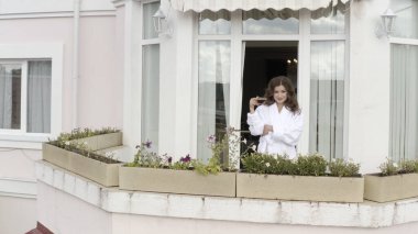 Beyaz bornoz giyen ve kırsal otelin balkonunda çay içen bir kadın. Video kaydı. Kadın sabahını bir fincan sıcak içecekle geçiriyor, rahatlıyor ve dışarı bakıyor..