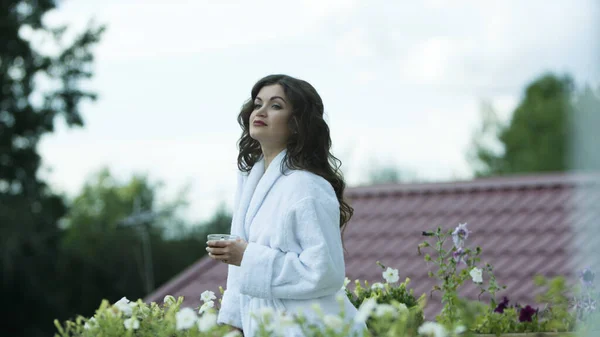 Mujer vestida con albornoz blanco y tomando té en el balcón del hotel rural. Vídeo. Mujer pasar la mañana con una taza de bebida caliente, relajarse y mirar a la distancia al aire libre. —  Fotos de Stock