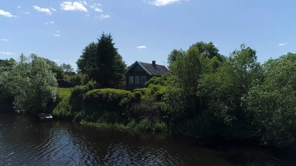 Sommer ländliche Landschaft mit Holzhaus in Seenähe. Schuss. Sonniger Tag in einem russischen Dorf mit einem kleinen Haus umgeben von Bäumen und Sträuchern auf blauem Himmel Hintergrund. — Stockfoto