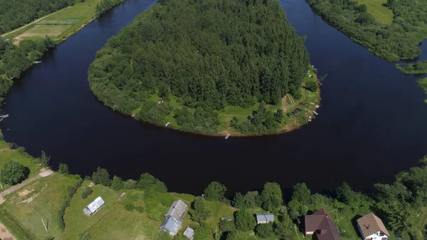 Flying over the wooden houses on the river shore. Shot. Aerial view of a small village and a curved narrow river bending around the green forested tiny island.