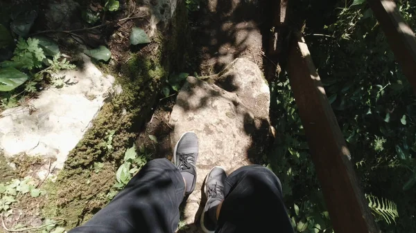 Erkek bacaklarının kırsal orman yolunda ilerleyişinin üst görüntüsü. Şarjör. Gri spor ayakkabı ve kot pantolon giyen bir adamı ormanda taş merdivenlerden inerken çeken ilk kişi..