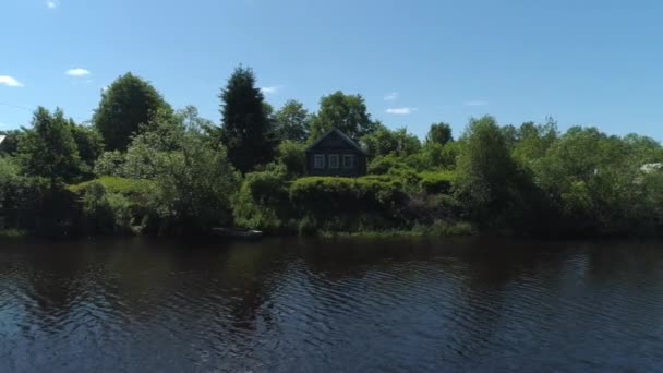 Veduta aerea di un fiume nel villaggio estivo e una vecchia casa in legno. Gli hanno sparato. Foresta e piccola casa circondata da alberi verdi e cespugli lungo la riva del fiume su sfondo cielo azzurro chiaro. — Video Stock