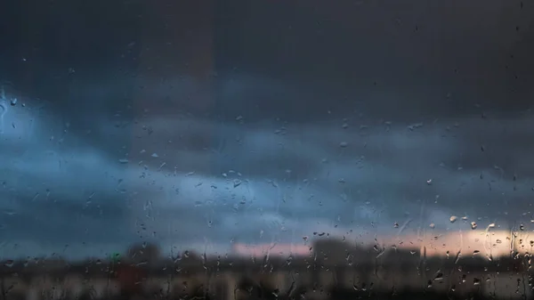 Deštivý den oknem na zamračené šedé obloze a na pozadí městských budov. Koncept. Večerní městská krajina za skleněným oknem s kapkami vody. — Stock fotografie
