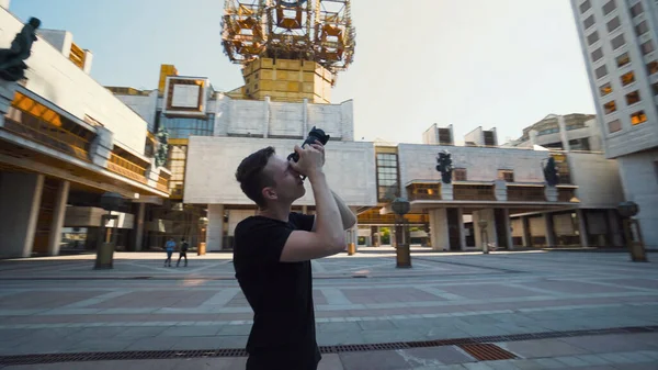 Jovem fotografa edifícios populares na cidade. Acção. Guy tira fotos profissionais da arquitetura urbana moderna. Fotógrafo tira fotos do grande edifício da cidade — Fotografia de Stock