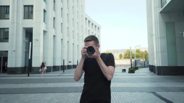 Professional photographer shoots you. Action. Young man takes you on professional camera while walking in city. Photographer walks around city with modern architecture — Stock Video