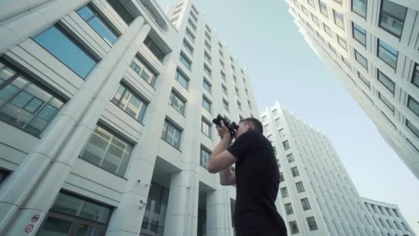 Young photographer takes picture of modern urban architecture. Action. Bottom view of guy photographing modern architecture with camera. Photographer shoots office buildings against blue sky — Stock Video