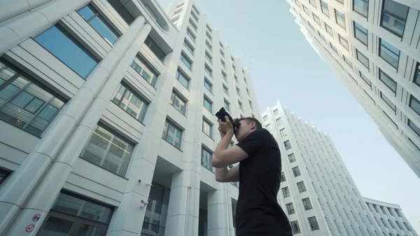 Молодий фотограф фотографує сучасну міську архітектуру. Дія. Вид знизу хлопця, який фотографує сучасну архітектуру з камерою. Фотограф застрелив офісні будівлі на тлі блакитного неба — стокове фото