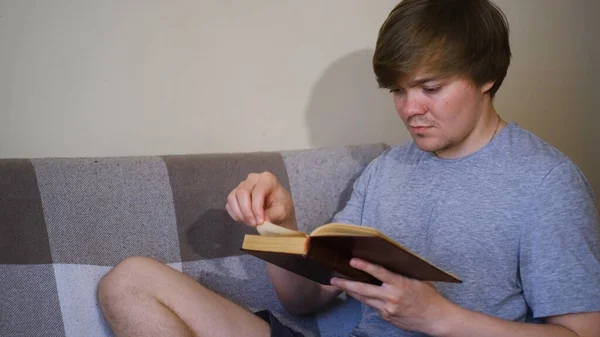 Junger Mann liest Buch und sitzt auf dem Sofa. Konzept. Student liest Klassiker mit angespanntem Blick. Mann liest altes Buch. Alte klassische Literatur — Stockfoto