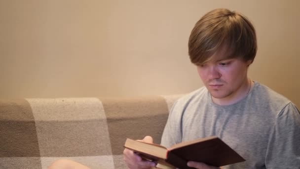 Young man reads book with interest. Concept. Student is intently reading old book sitting on sofa. Erudite young man reads book in evening. Reading books is good for brain — Stock Video