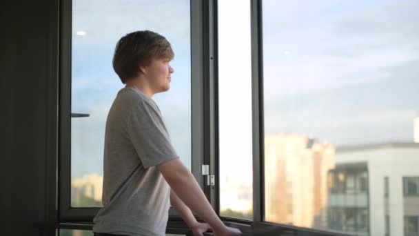 Młody człowiek cieszy się widokiem z okna stojącego na balkonie. Koncepcja. Student z radością obserwuje życie za oknem na tle nieba. Student stoi na balkonie i wygląda przez okno — Wideo stockowe