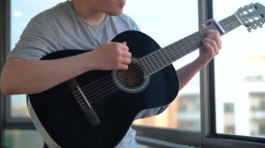 Pencere arkasında gitar çalan adamın yakın çekimi. Kavram. Adam güzel siyah gitar çalıyor. Profesyonel gitarist pencereden arka planda gökyüzünde çalar