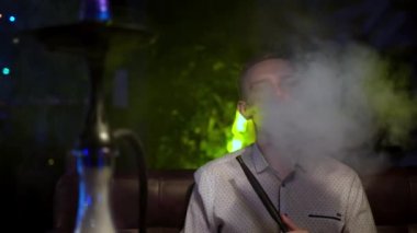 Bekar adam nargile içiyor. Medya. Düşünceli adam nargile dumanını neon lambanın arkasına üfledi. İnsan dinlenir ve nargile üzerine kafa yorar.