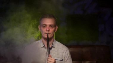Bekar adam nargile içiyor. Medya. Düşünceli adam nargile dumanını neon lambanın arkasına üfledi. İnsan dinlenir ve nargile üzerine kafa yorar.