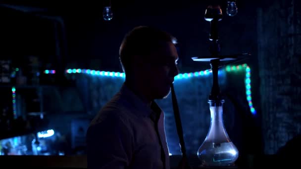 Αρσενική σιλουέτα που καπνίζει ναργιλέ στο μπαρ. Μέσα ενημέρωσης. Ο άνθρωπος καπνίζει ναργιλέ στο μπαρ στο σκοτάδι στο φόντο του μπλε φωτός. Νυχτερινό μπαρ με ναργιλέ — Αρχείο Βίντεο