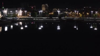 Gece şehrin ışıklarını yansıtan karanlık su manzarası. Stok görüntüleri. Yürüyen insanların ve gece parlayan ışıkların olduğu güzel şehir barajı. Şehrin gece hayatı nehir suyuna yansıyor.
