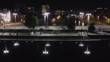İnsanlar geceleri şehirde gezinti güvertesinde yürüyorlar. Stok görüntüleri. Modern şehrin merkezindeki yoğun köprünün gece manzarası. Akşamın ışıkları karanlık sulara yansıyor..