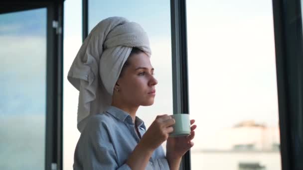 Kobieta z ręcznikiem na głowie pijąca kawę w tle okna. Koncepcja. Piękna młoda kobieta przygotowująca się do rozpoczęcia dnia pracy filiżanką kawy na balkonie — Wideo stockowe