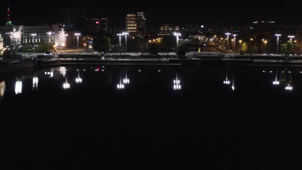 Vista superior del agua oscura reflejando las luces de la ciudad por la noche. Imágenes de archivo. Hermosa presa de la ciudad con gente caminando y luces brillantes por la noche. La vida nocturna de la ciudad reflejada en el agua del río — Vídeos de Stock