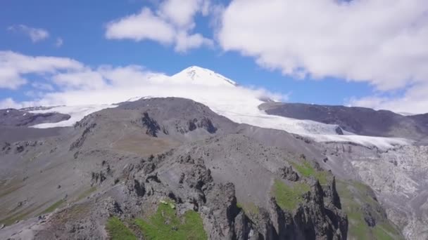 Paisagem de montanha com pico nevado e sopé rochoso. Clipe. Vista deslumbrante da montanha nevada em nuvens no fundo do céu azul. Trilhas de montanha em lugares rochosos levam ao pico nevado — Vídeo de Stock