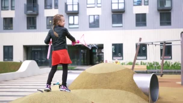 Cute girl gra z gigantycznych baniek mydlanych w pobliżu domu. Akcja. Biała kobieta dziecko dmuchanie duże pęcherzyki podczas stania na dużym sztucznym wzgórzu na placu zabaw. — Wideo stockowe