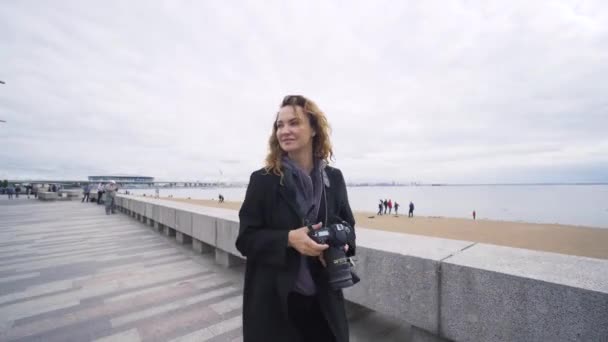 La fotógrafa camina por el terraplén. Acción. Mujer toma fotos de terraplén con el paisaje marino en la cámara. Mujer de vacaciones toma una foto de terraplén en el mar de fondo en tiempo nublado — Vídeos de Stock