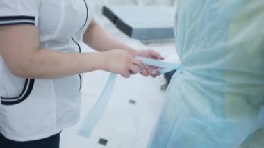 Hemşire, cerrahlara kıyafet konusunda yardım ediyor. Başla. Cerrahların bornozlarını bağlamaya yardım eden hemşirelerin yakın çekimi. Cerrah yardımcısı ameliyat için bornoz giymene yardım eder.