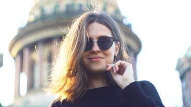 Katedral arka planında, şehir sokaklarında gülümseyen güneş gözlüğü takan güzel bir kız. Medya. Siyah örgü süveter giyen, gözlüğünü takan ve ayakta gülümseyen seksi genç bir kadının portresi.